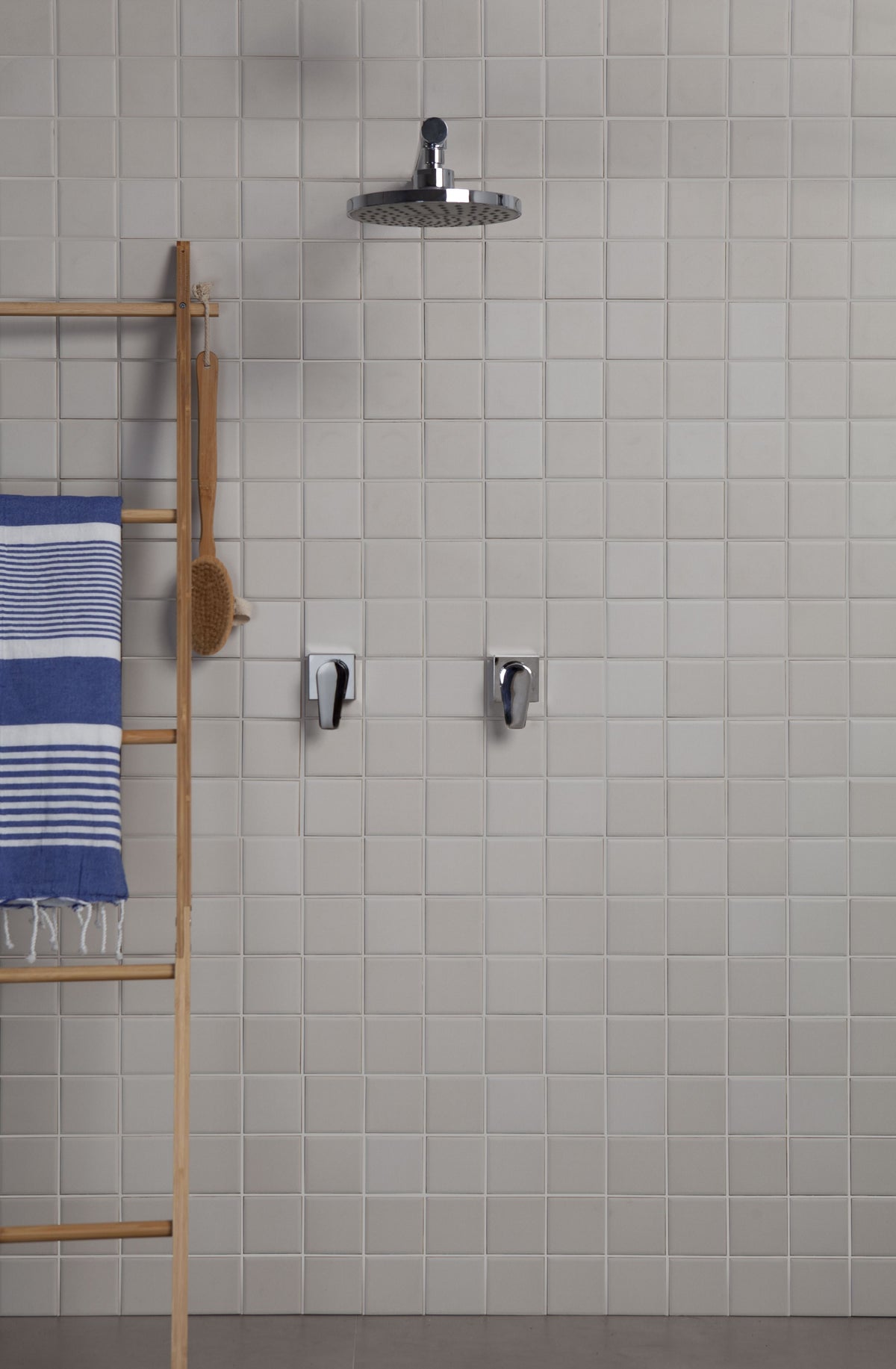 A modern wall-mounted rain shower head with chrome fixtures installed on a light gray tiled bathroom wall, with a wooden towel rack and blue striped towel visible on the left.
