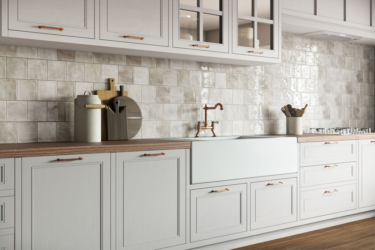 A modern kitchen interior with white shaker-style cabinets, a glossy light gray tile backsplash, wood countertops, and a white farmhouse sink with a copper faucet.