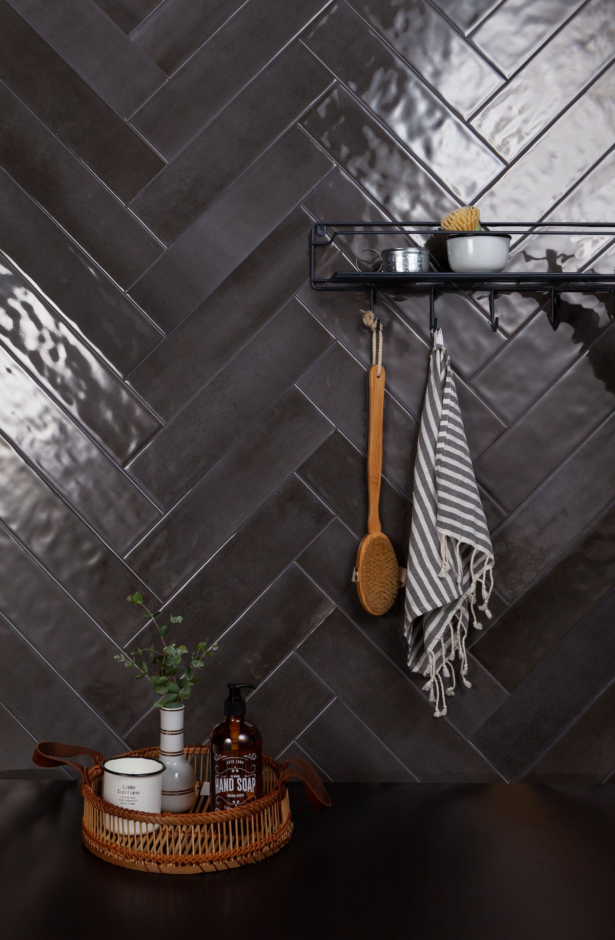 Dark charcoal gray herringbone subway wall tiles in a modern kitchen setting, featuring a black floating shelf with small bowls, a wooden spoon, and a striped towel.