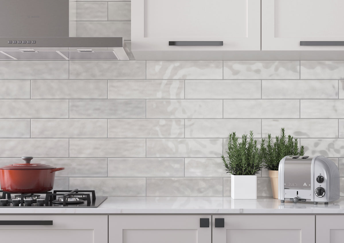 Light gray subway tile backsplash installed on a kitchen wall, shown with white cabinets, a gas stove with a red pot, potted green plants, and a silver toaster.