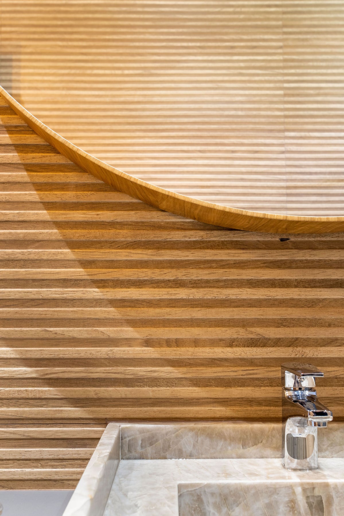 A close-up of a modern bathroom sink with a chrome faucet set into a light stone basin, set against a curved wooden slat wall.