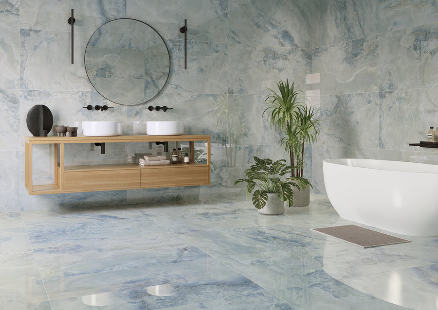 A modern bathroom interior with blue-gray marble-look tiles, a floating light wood double-sink vanity, a round wall mirror, a freestanding white bathtub, and potted green plants.