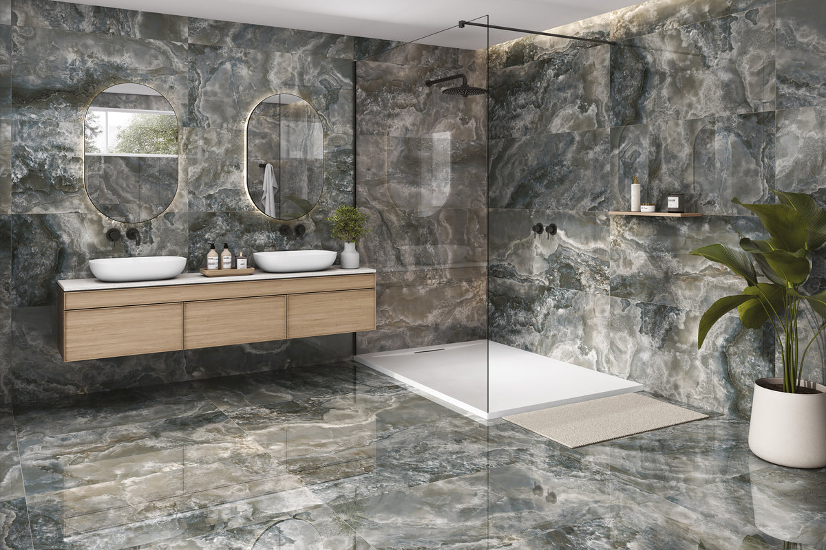 A modern bathroom interior with large black marbled stone look porcelain tiles, a double-sink vanity with light wood cabinets, oval mirrors, a glass shower enclosure, and a potted plant.