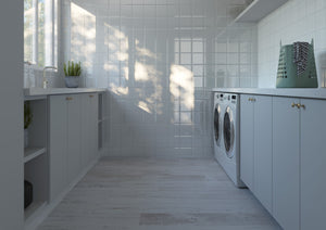 A bright, modern laundry room with white cabinets, white tiled walls, a tiled back wall, and a washer and dryer built into the cabinetry. Sunlight streams in from a window on the left, casting shadows on the wall and floor.