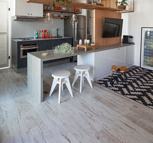 A modern kitchen island with a light gray concrete-look top and white pedestal base, paired with two white stools. The background shows a dark gray kitchen with stainless steel appliances, open wood shelving, and a light gray wood-look floor.