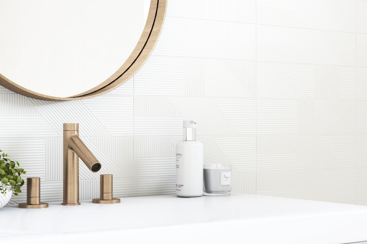 A modern bathroom scene featuring a matte brass faucet with matching handles on a white countertop, set against a white textured tile wall with a round mirror partially visible at the top.