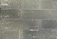 Close-up of Artesano Tulum Sage 2x9 glossy clay brick tile in running bond, rippled sage-green glaze with red clay chips